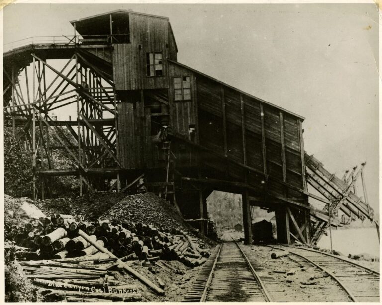 Large wooden coal bunker on pilings spanning two railroad tracks beside a lake