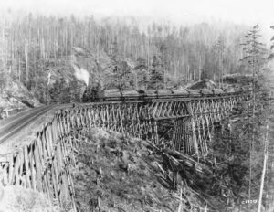 A loaded log train crossing a tall wooden railroad trestle in a forested river valley, 1926