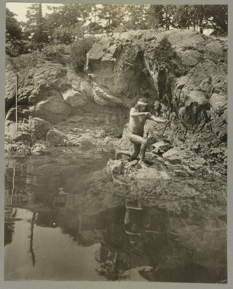 1924 photograph of a Lummi man kneeling on rocks beside still water, drawing a bow and arrow to shoot a fish