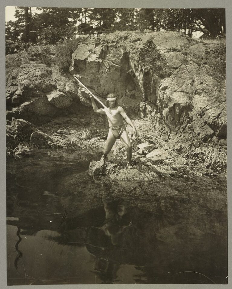 1924 photograph of a Lummi man standing on rocks beside a tidal pool, holding a long fishing spear raised over the water