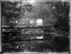 1902 photograph of Whatcom Falls with a log bridge crossing above the waterfall